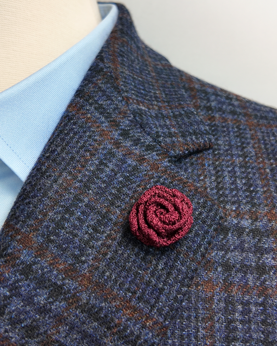 A close-up of a textured burgundy rose-shaped brooch lapel flower pinned to a blue and brown plaid tweed jacket, worn over a light blue dress shirt.