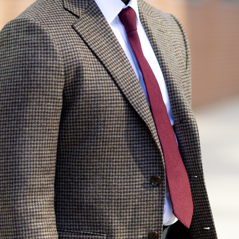 A close-up of a man wearing a brown and black houndstooth blazer, a white dress shirt, and a burgundy tie.