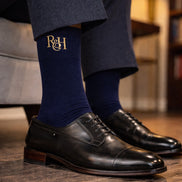 Person wearing navy socks with gold logo and black dress shoes on a wooden floor.