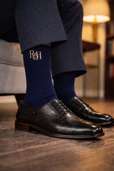 Person wearing navy socks with gold logo and black dress shoes on a wooden floor.