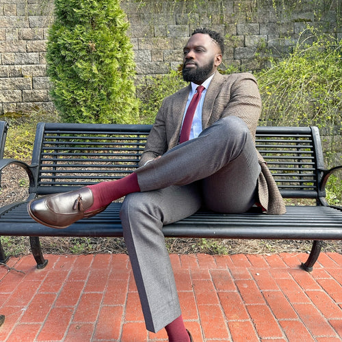 Man in a suit sitting on a bench outdoors with a stone wall and greenery in the background