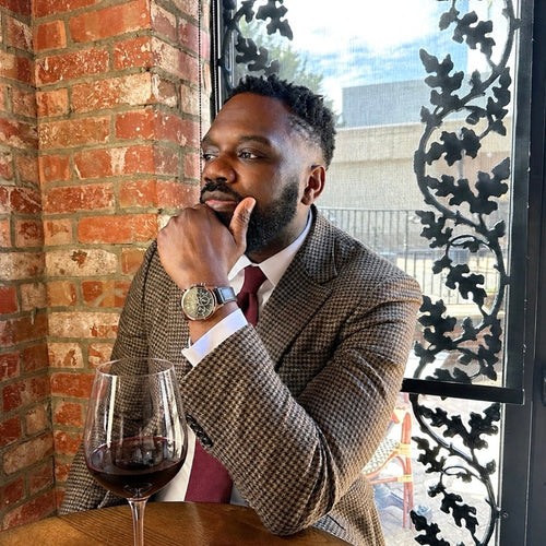 Man in a suit with a glass of red wine leaning against a brick wall.