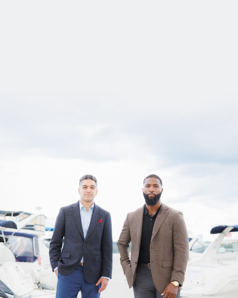 Two men standing outdoors in front of a bright overcast sky, with boats visible in the background. The man on the left wears a dark blue patterned blazer and blue trousers, and the man on the right wears a brown blazer over a black shirt with gray trousers.