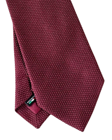 A close-up of a textured wine red necktie with a small green and white brand tag near the tip, set against a plain white background.