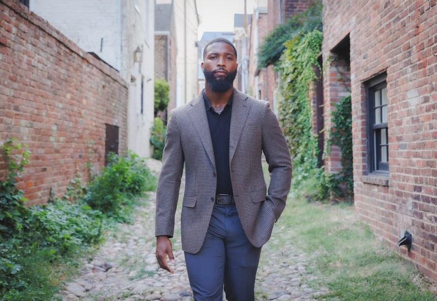 A man wearing a textured gray blazer, black shirt, and blue trousers walking down a cobblestone alleyway lined with brick buildings and green ivy.