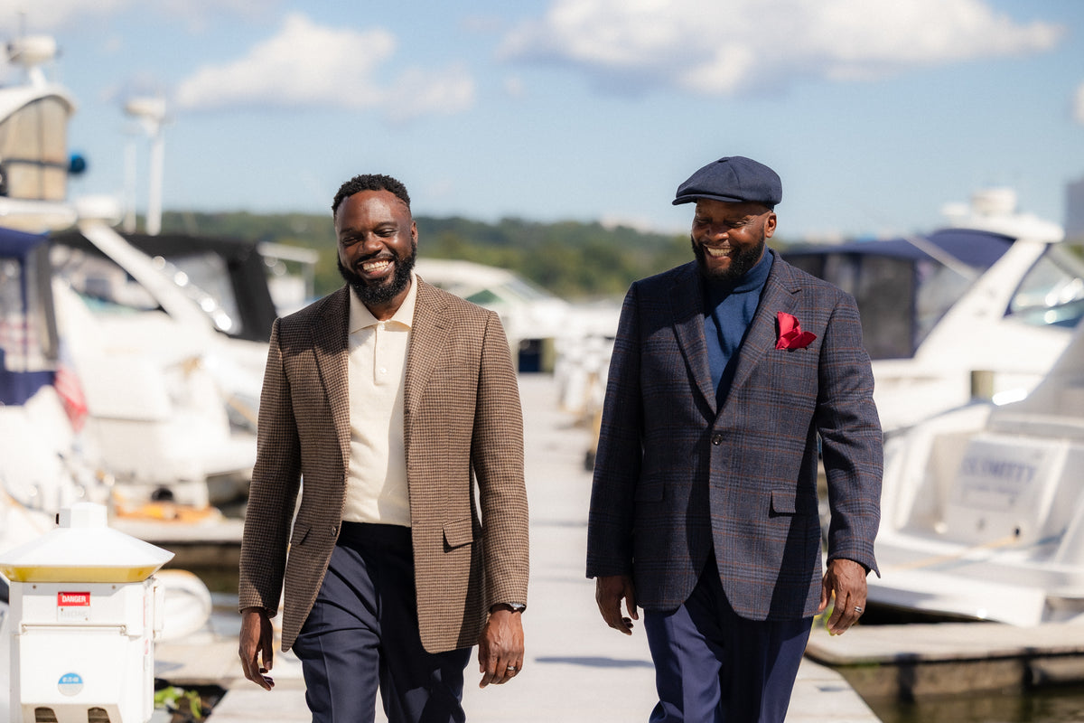 Two men walking side by side on a docked boatyard path. The man on the left wears a brown blazer over a cream shirt and navy pants.