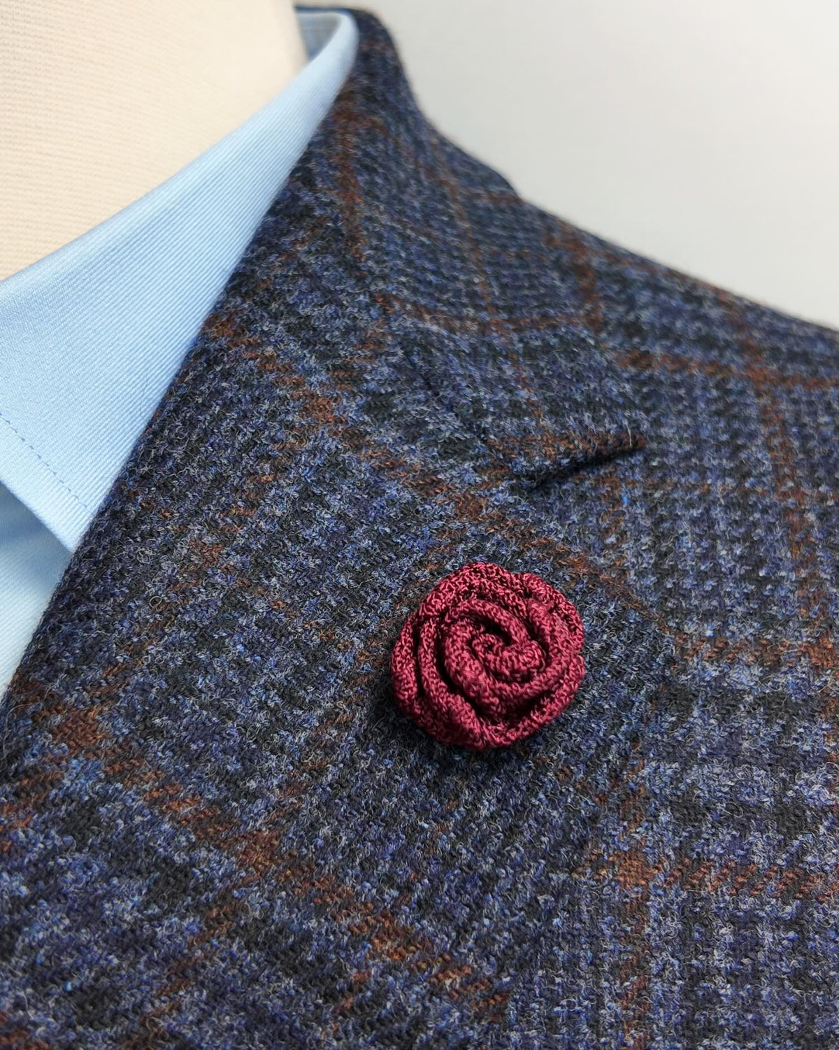 A close-up of a textured burgundy rose-shaped brooch pinned to a blue and brown plaid tweed jacket, worn over a light blue dress shirt.