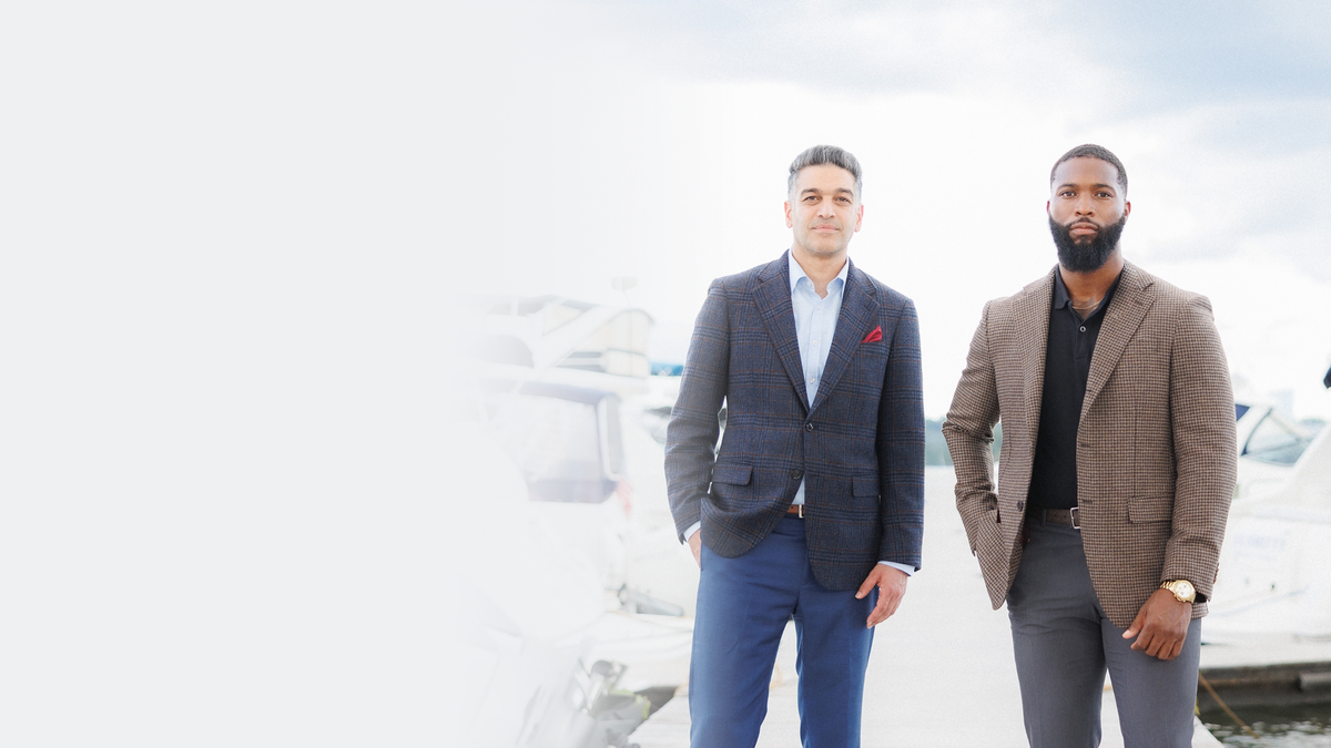 Two men standing outdoors near a dock or marina, wearing tailored blazers. The man on the left wears a dark charcoal plaid blazer with blue trousers, and the man on the right wears a taupe-brown blazer with gray trousers.